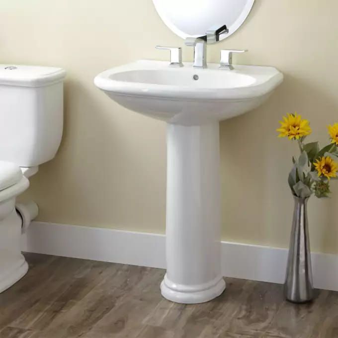 White pedestal sink and toilet in a bathroom, flowers in a vase, wood-look floor.