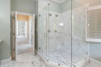Bathroom with glass shower, marble walls, and pebble floor; open door leads to hallway with carpet.