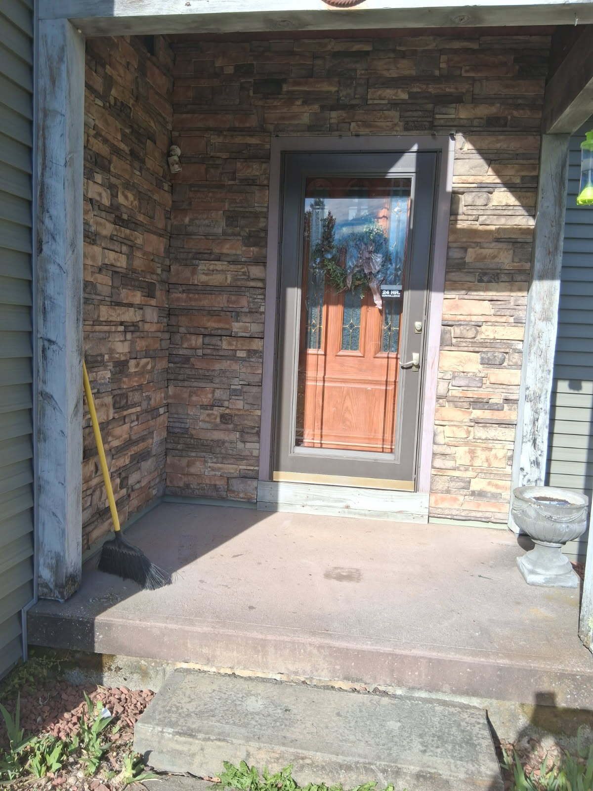 Entryway with stone veneer wall, concrete porch, and wooden door. A broom leans against a support column.