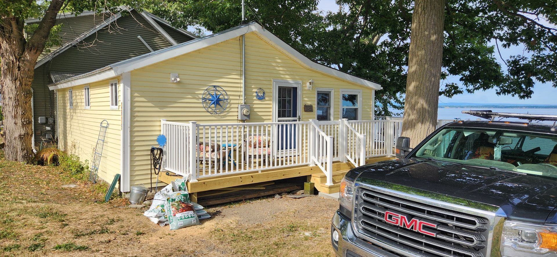 Yellow cottage with a white porch. A black GMC truck is parked nearby, with a view of the water in the background.