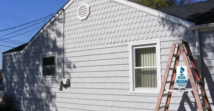 Gray shingled house with a window and a ladder propped against it, on a sunny day.