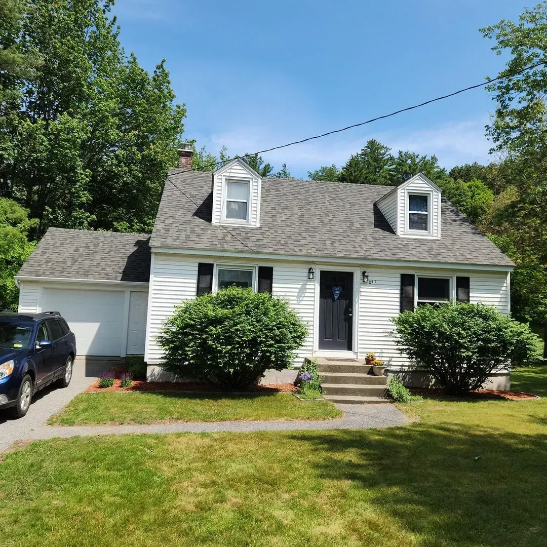 White house with black door, garage, dormer windows, shrubs, and blue SUV.