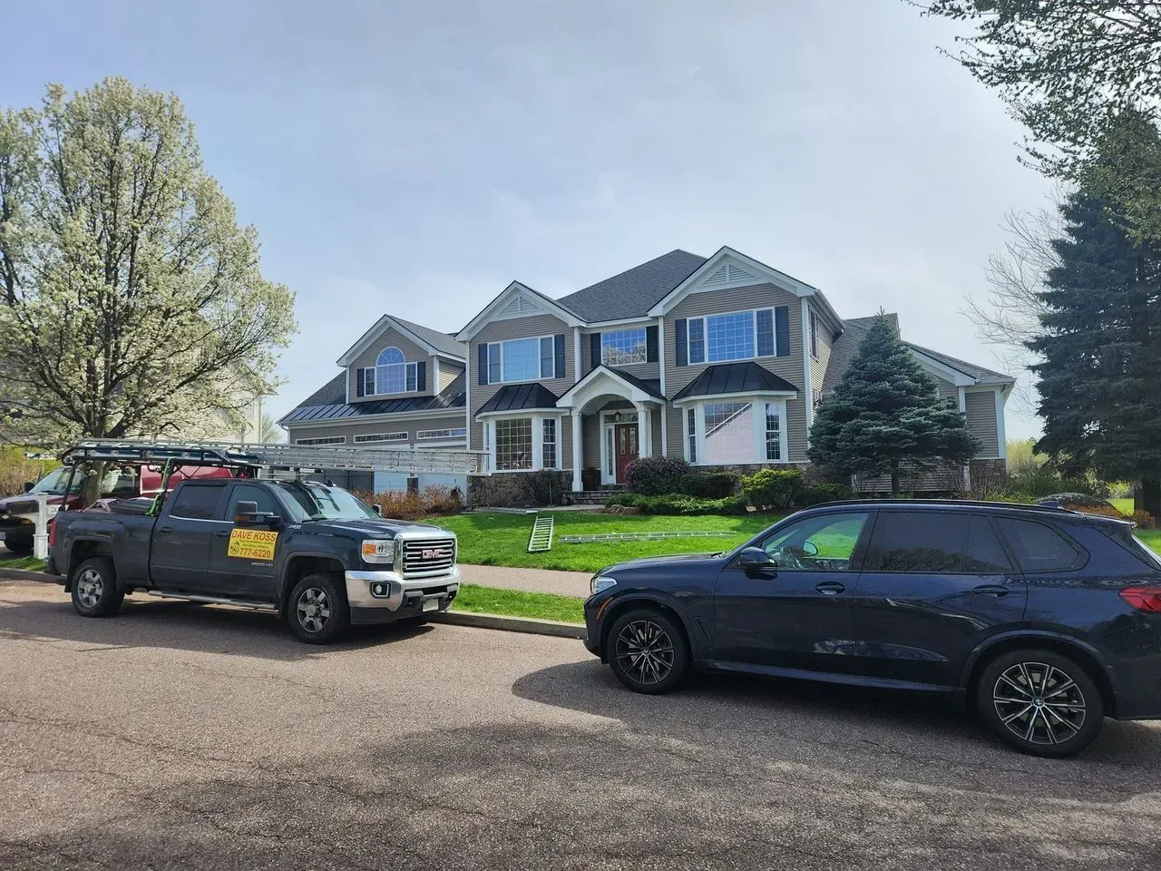 House with dark roof, truck and SUV parked on street. Light blue sky and blossoming tree.