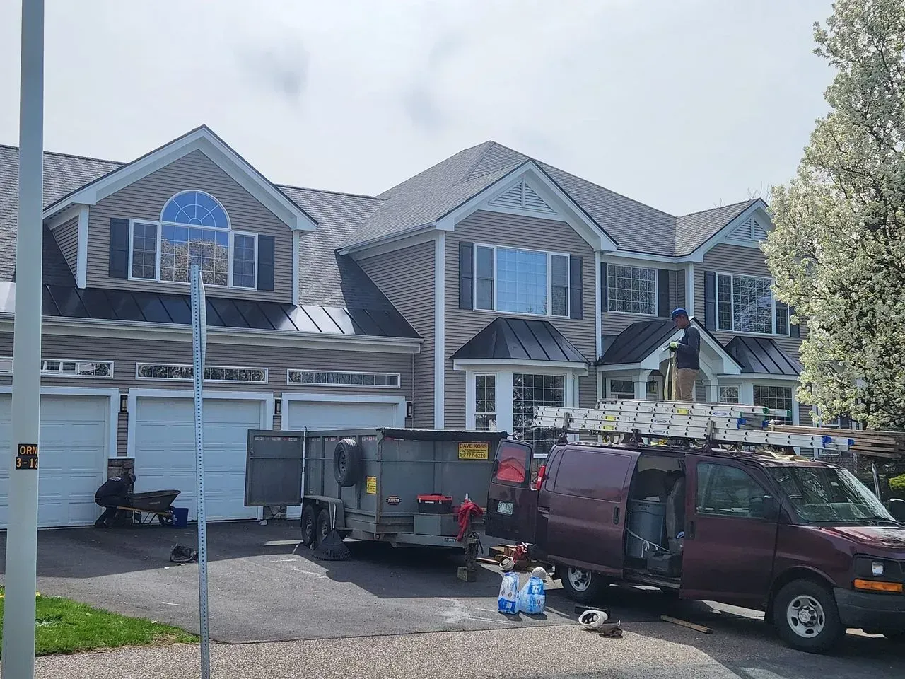House with roofing materials, van, and trailer parked outside. Roof work in progress.
