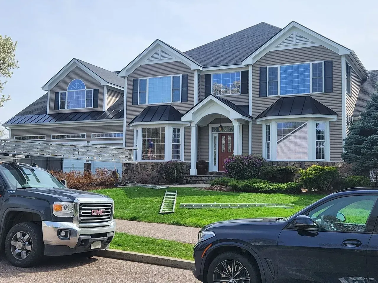 Two-story house with tan siding, black shutters, and dark roof; two vehicles parked in front.