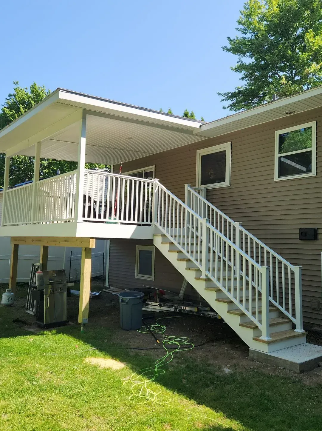 A covered wooden deck with white railing, stairs, and siding, attached to a brown house with a blue sky background.