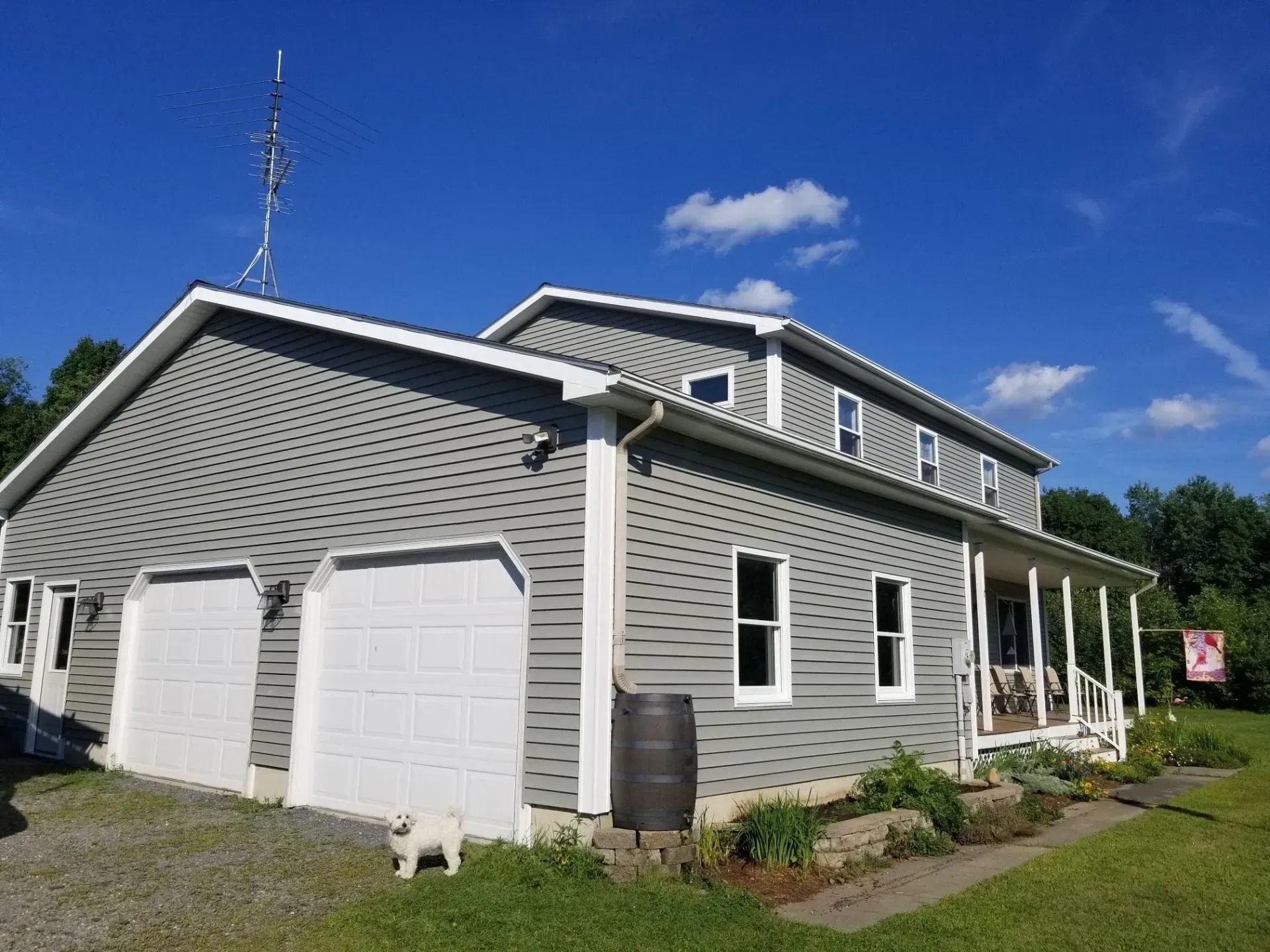 Two-story gray house with white trim and garage doors, a small dog, and a blue sky.