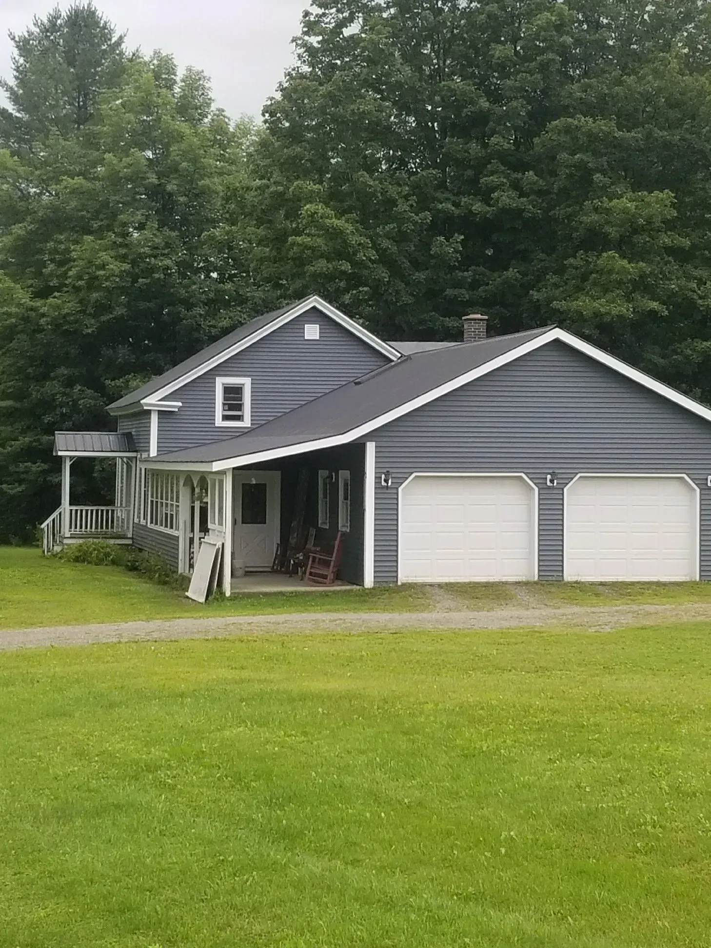 Blue house with white garage doors, porch, and green lawn. Tall trees in background.