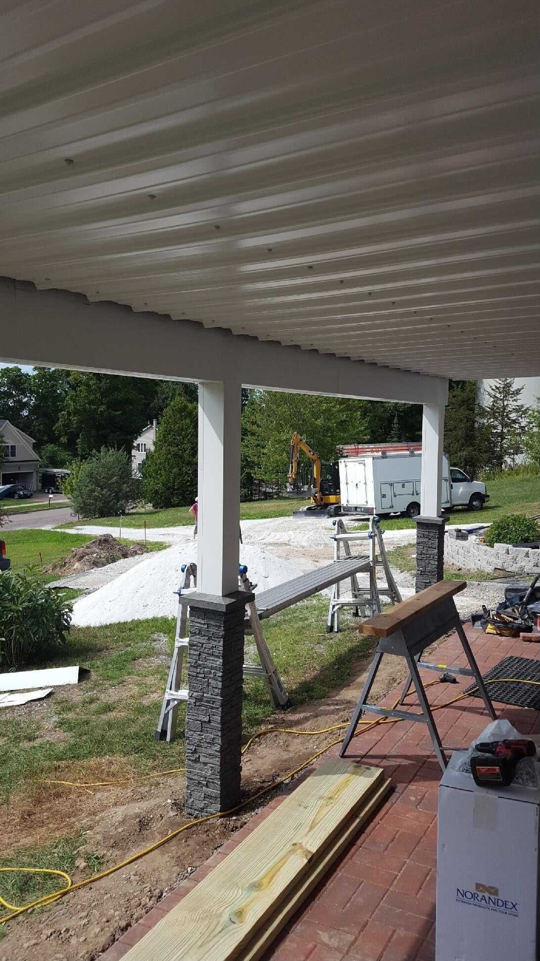 Porch under construction with white columns, stone accents, and lumber; view of a yard with construction equipment.