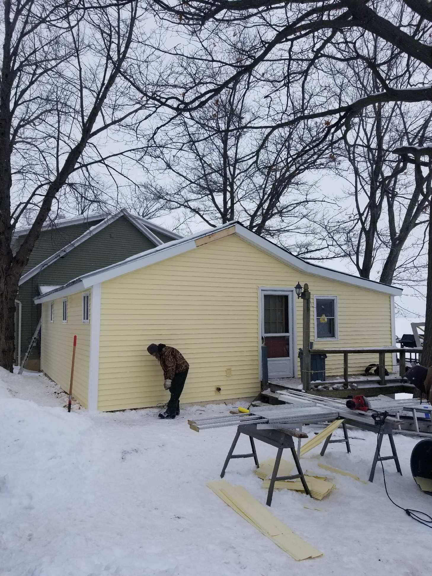 Construction worker outside a yellow house in the snow. Tools, wood planks, and snow surround him.