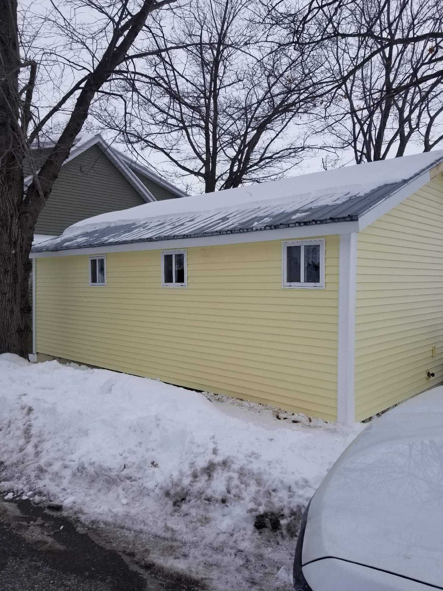 Yellow building with three windows, snow on the ground and roof, next to bare trees.