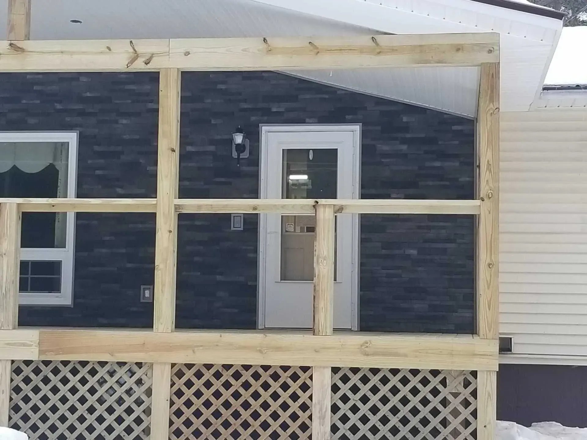 Wooden deck with lattice panels and railing, in front of a house with a dark stone facade and white door.