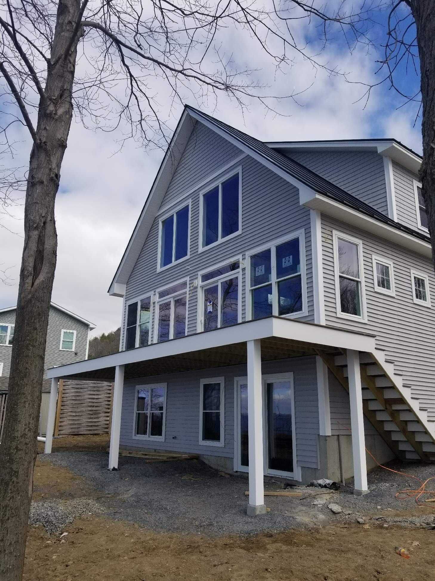 Gray two-story house with large windows, covered deck, and stairs. Trees frame the house against a cloudy sky.