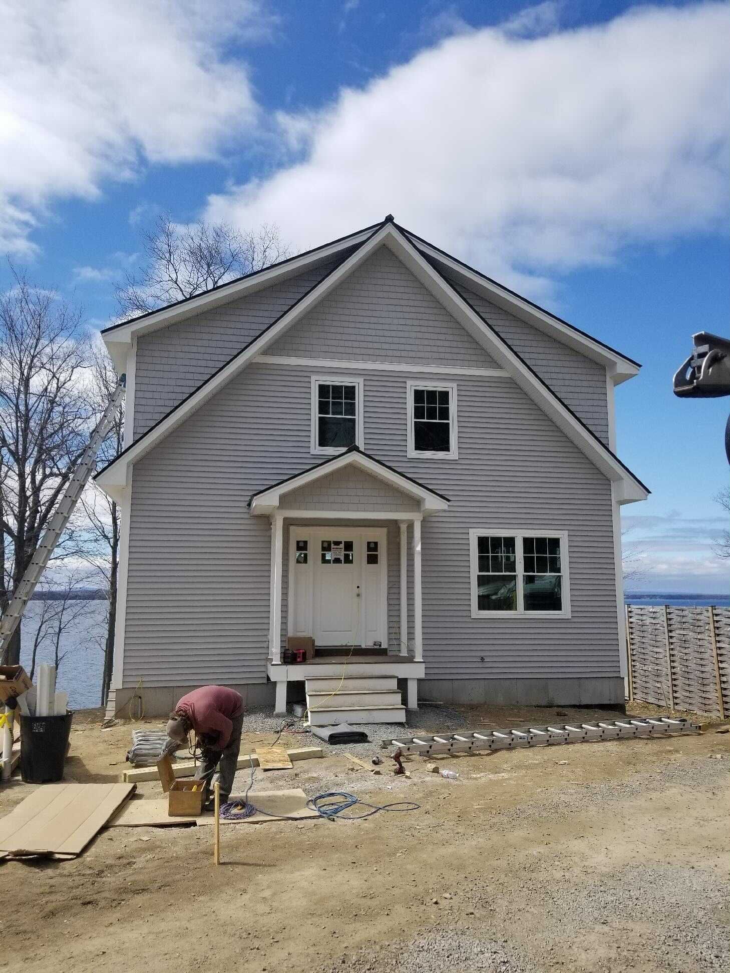 Two-story house with gray siding under a cloudy sky. A person works on the landscaping in front of the house.
