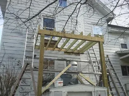 Wooden deck frame construction attached to the side of a white house with multiple ladders and supports.