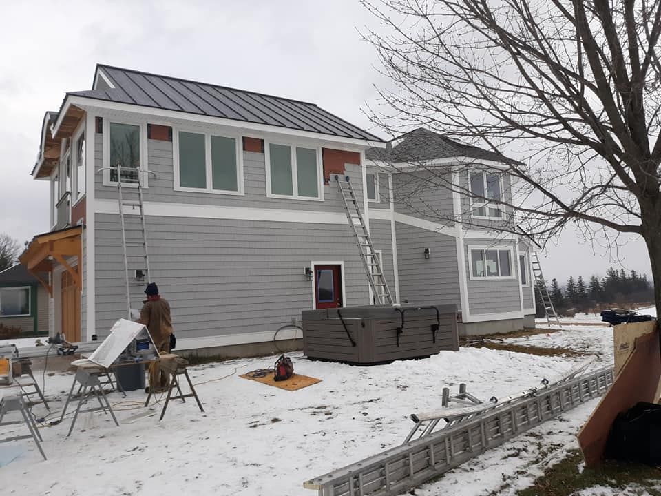 A house under construction in a snowy setting with a worker on a ladder, various tools, and gray siding.