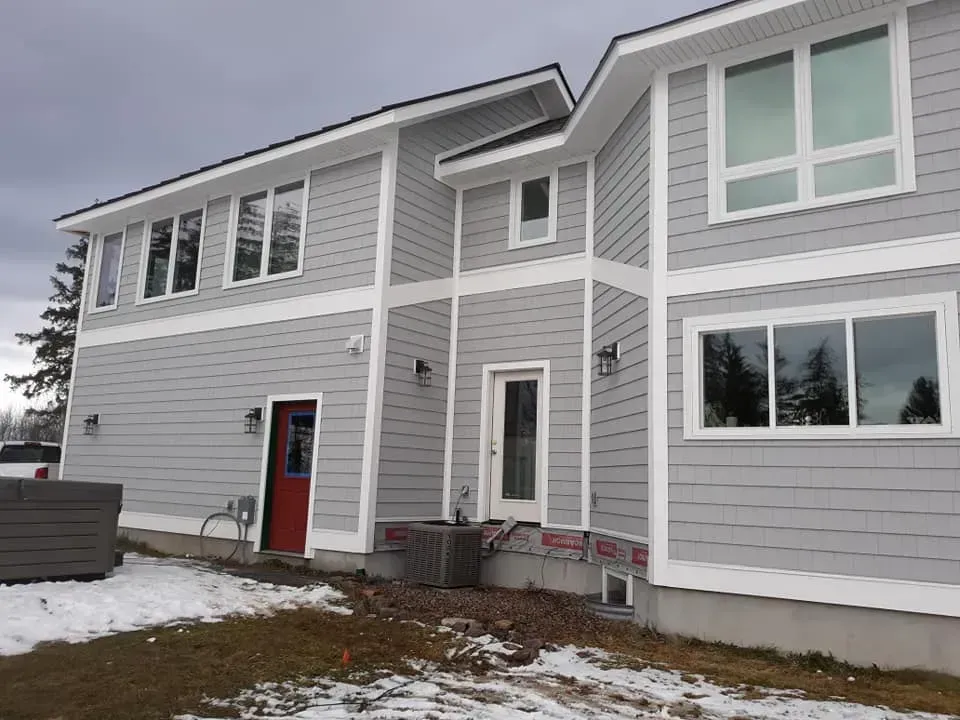 Gray house with white trim and a red door, set in a snowy, overcast environment.
