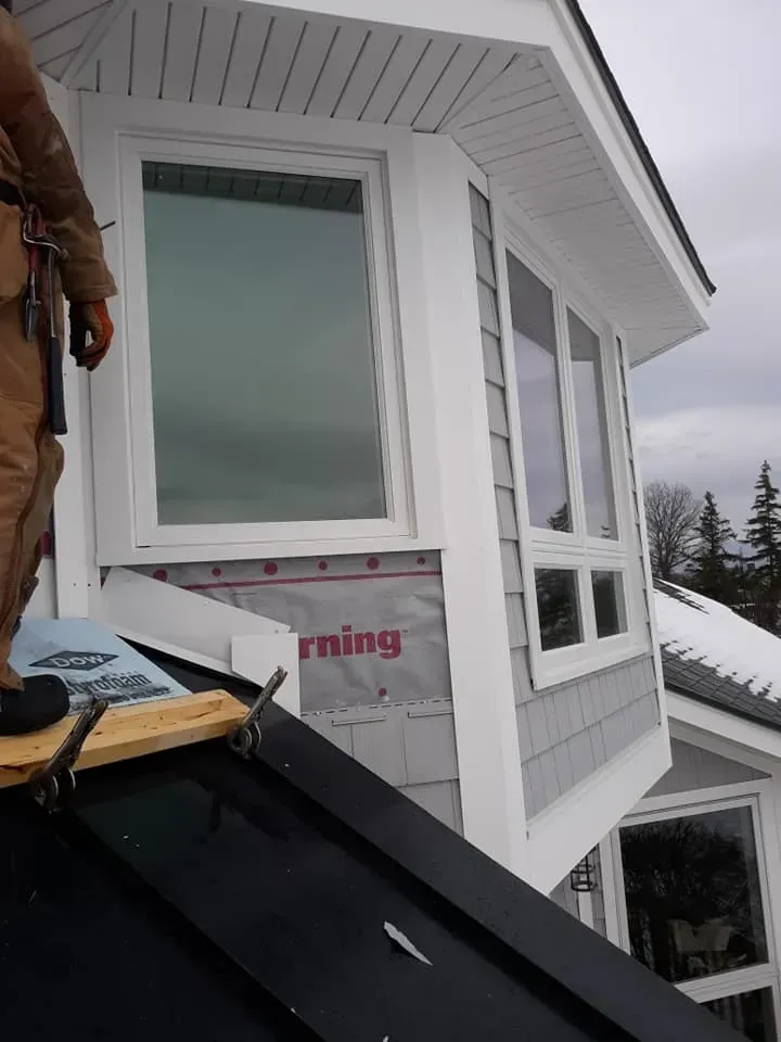Person on rooftop near bay window with gray siding. Construction in progress.