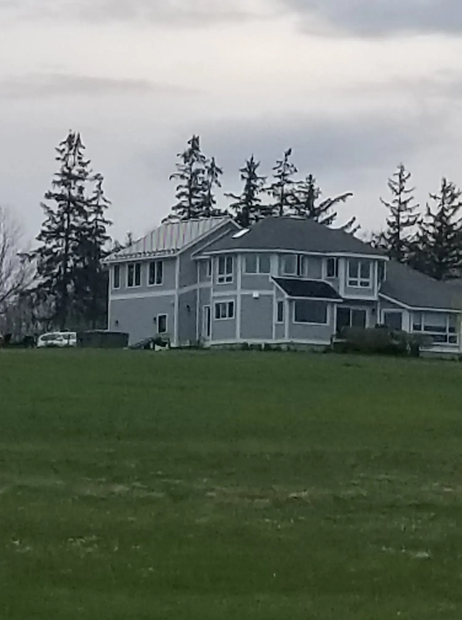 A light grey two-story house with a green lawn, trees, and an overcast sky.
