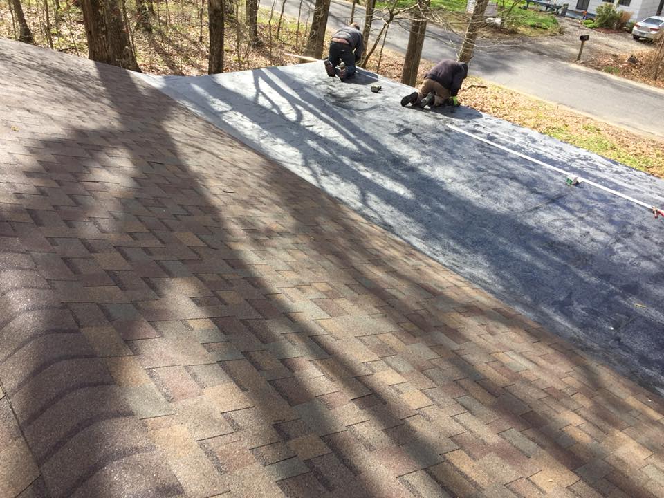 Roofers installing shingles on a roof, with trees and a street in the background.