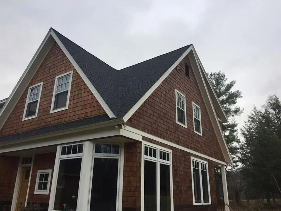 Brown shingled house with white trim and dark roof under a cloudy sky.
