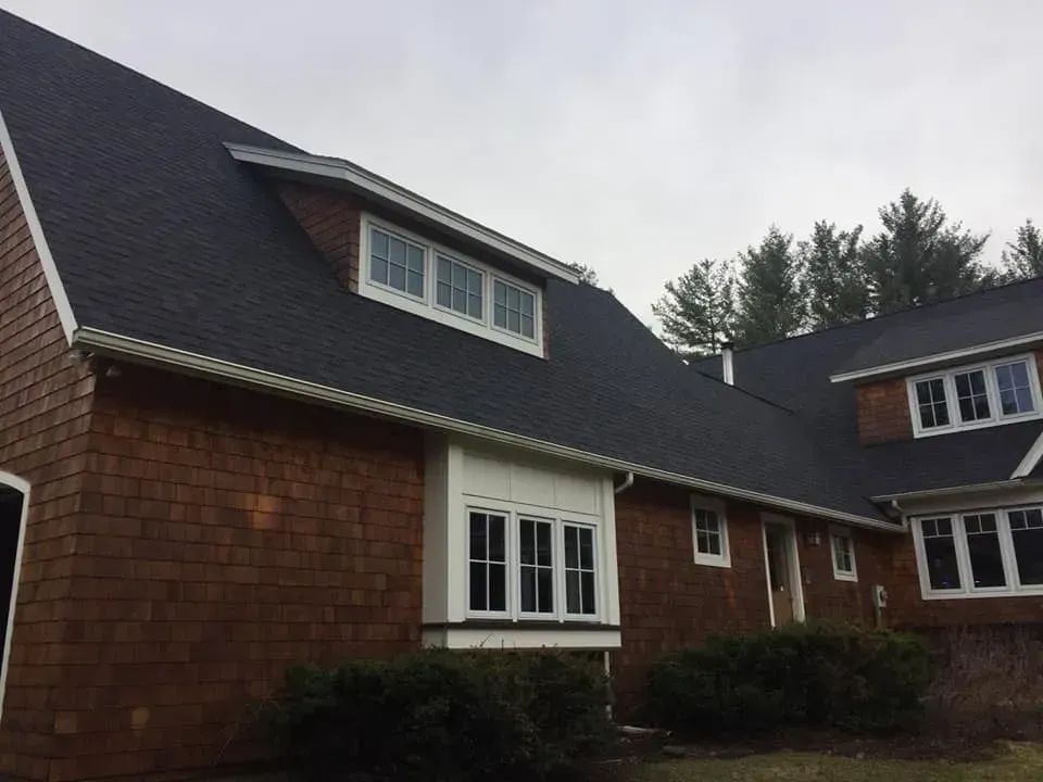 Brown shingled house with dark roof and white trim under an overcast sky.