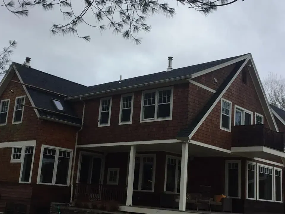 Brown shingle house with a dark roof and white trim under an overcast sky.