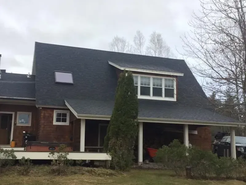 Brown house with a dark roof and a porch, set against a cloudy sky.