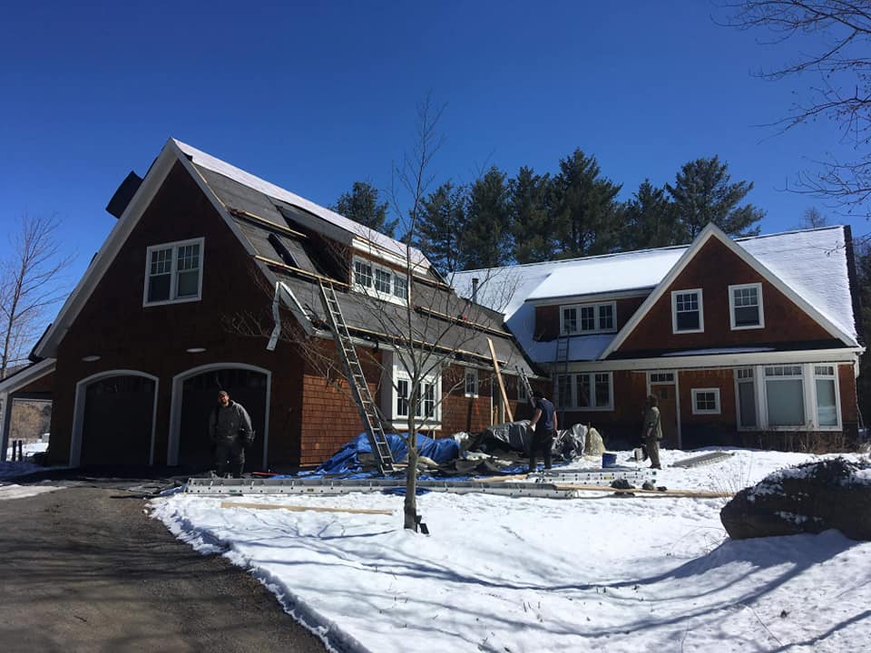 House under construction; workers on the roof in the snow. Red siding, blue sky, garage visible.