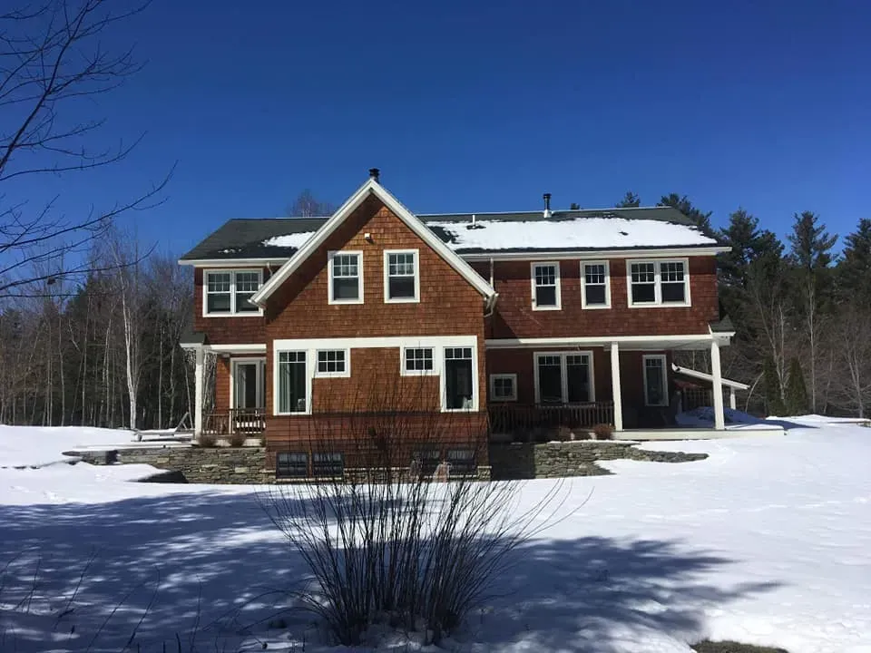 Two-story brown shingle house with white-framed windows, surrounded by snow, under a clear blue sky.