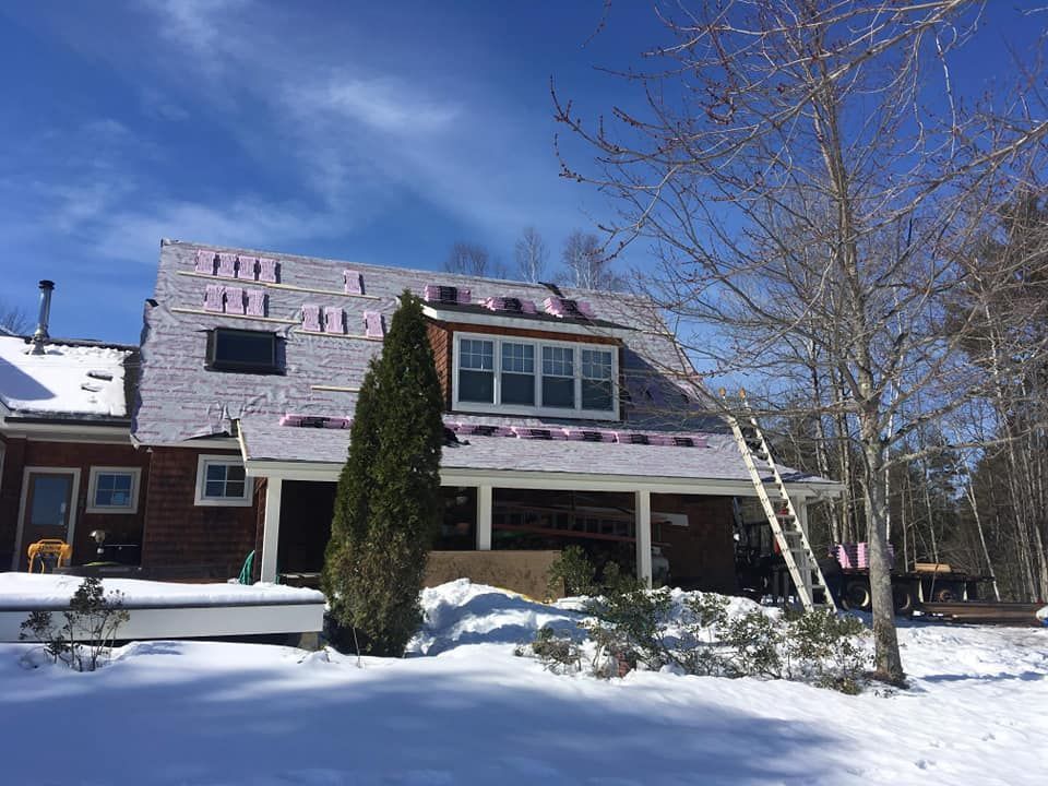 House with a partially replaced roof; snow on the ground, ladder against the side, clear sky.