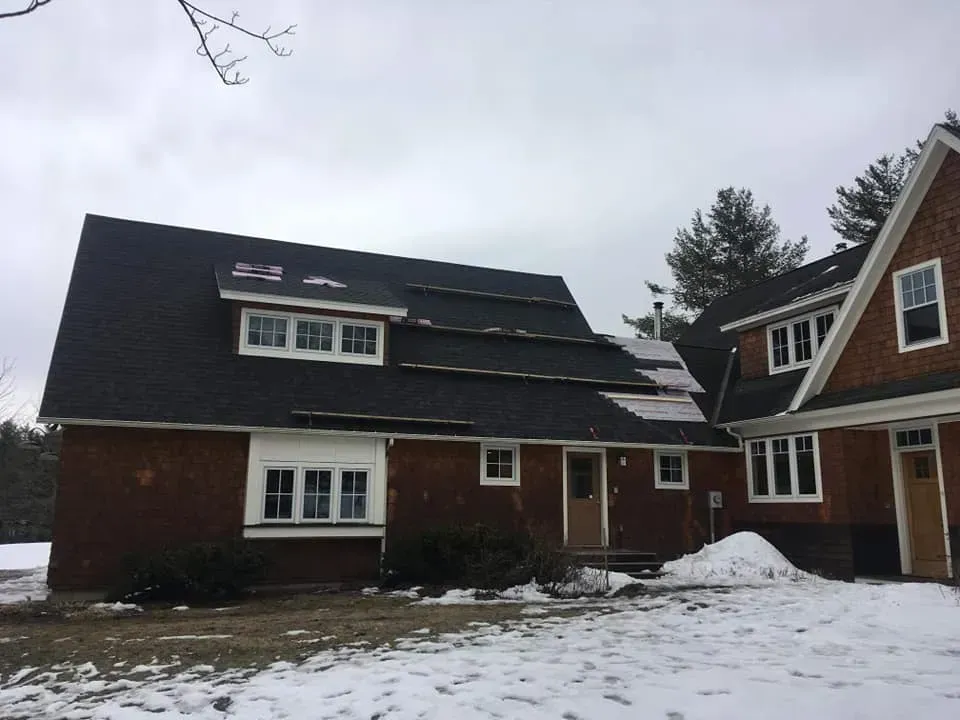Red brick house with a dark roof covered with patches of snow under a cloudy sky.
