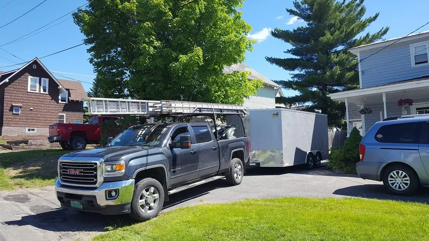 Truck pulling a trailer in a driveway; ladder on the truck's roof.