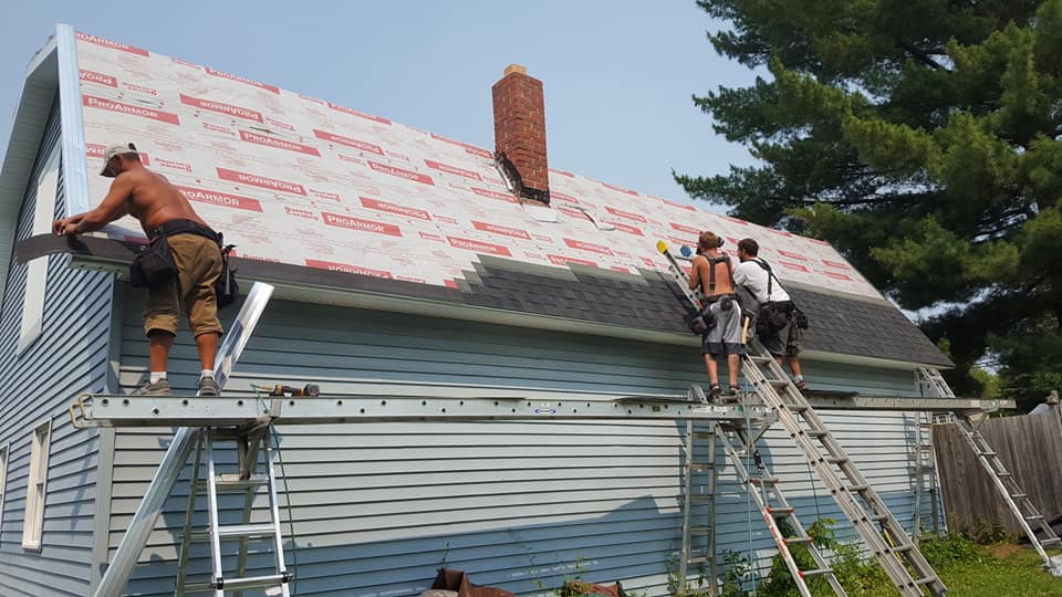 Roofers working on a light blue house, replacing shingles. Ladders and scaffolding are in use.