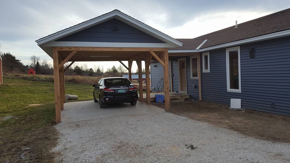 Car parked under a wooden carport attached to a blue house with a gravel driveway.