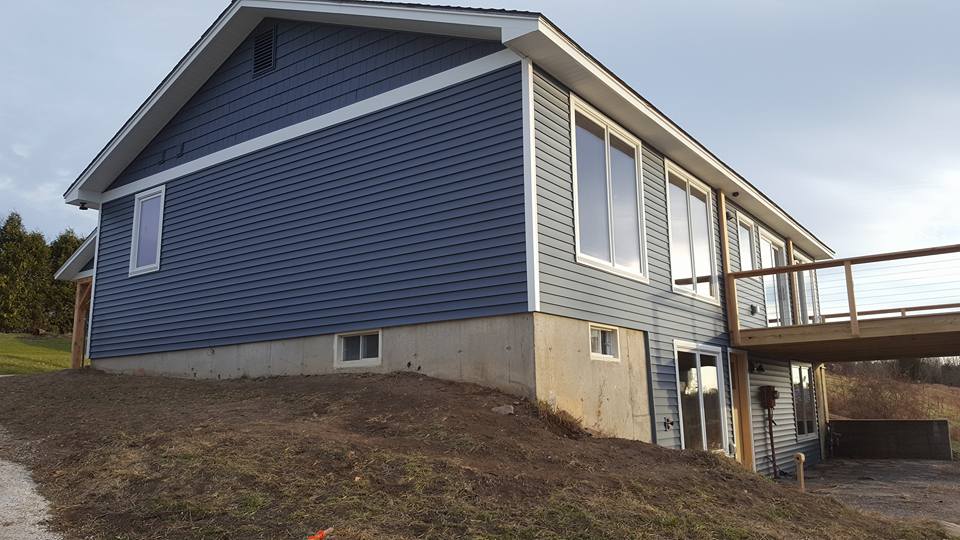 Blue-sided house with a deck and large windows on a grassy hill.