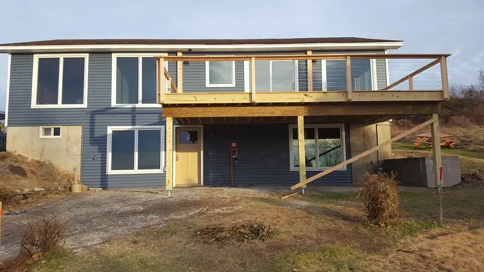 Two-story blue house with wooden deck, set on gravel, sunny day.