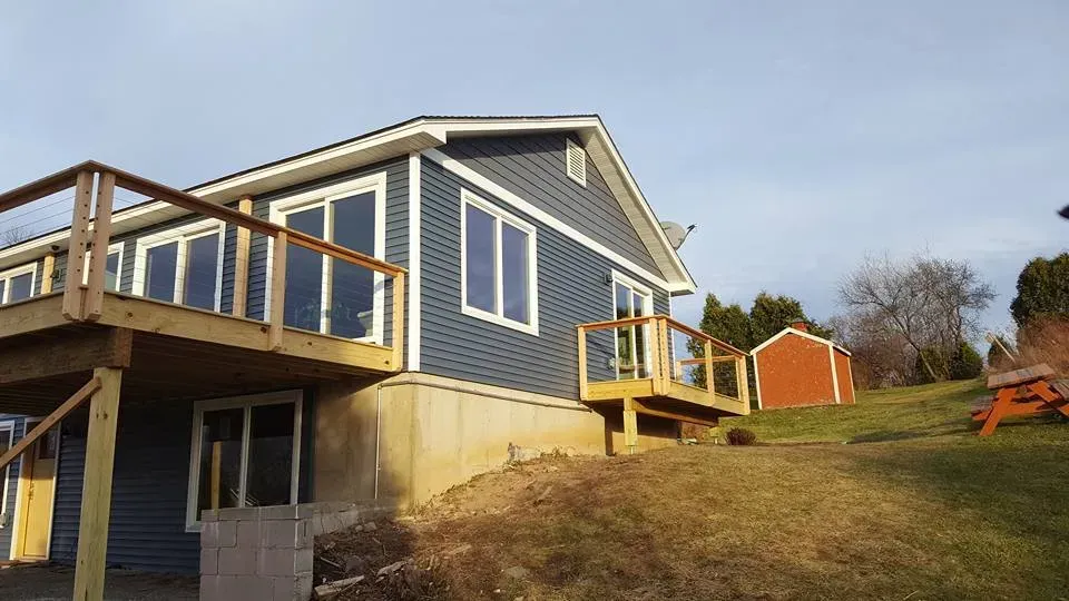 Blue-sided house with wooden deck, set on a grassy hill; small red shed in the background.