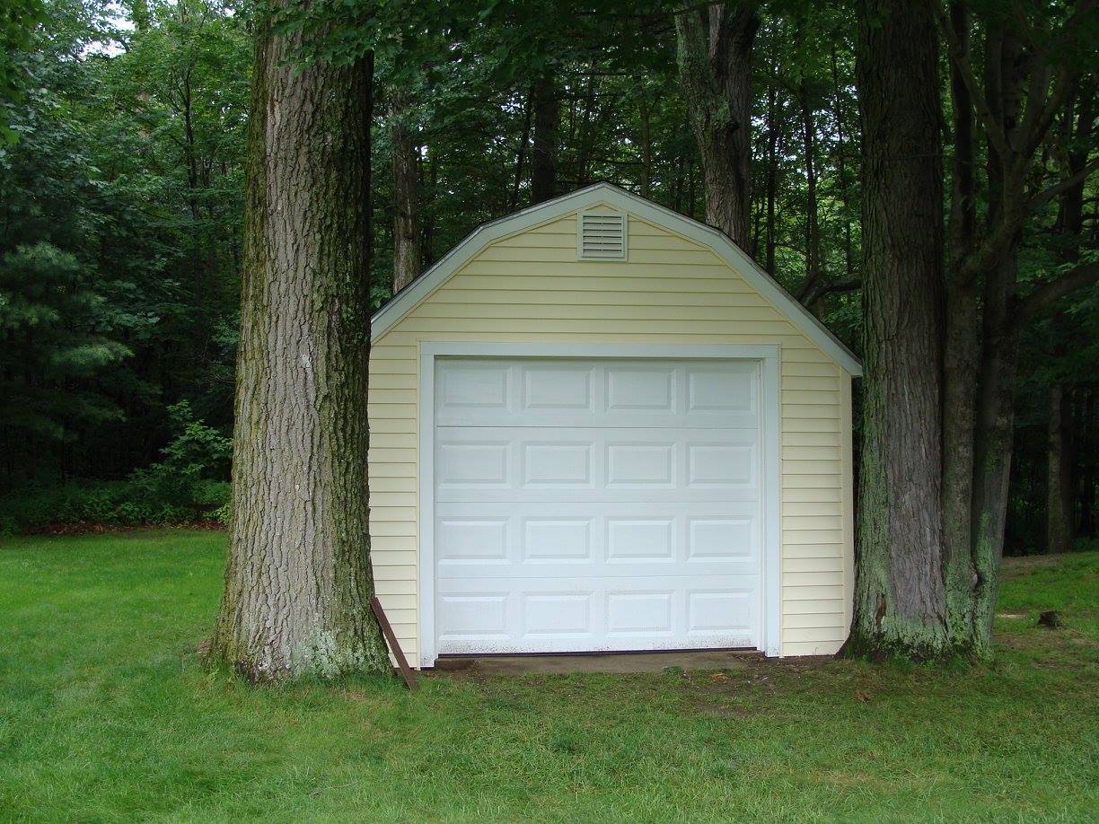 Yellow garage with white door nestled between two trees on a grassy lawn.