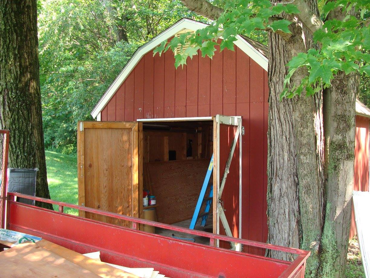 Red shed with open wooden doors, surrounded by trees and a red structure in the foreground.