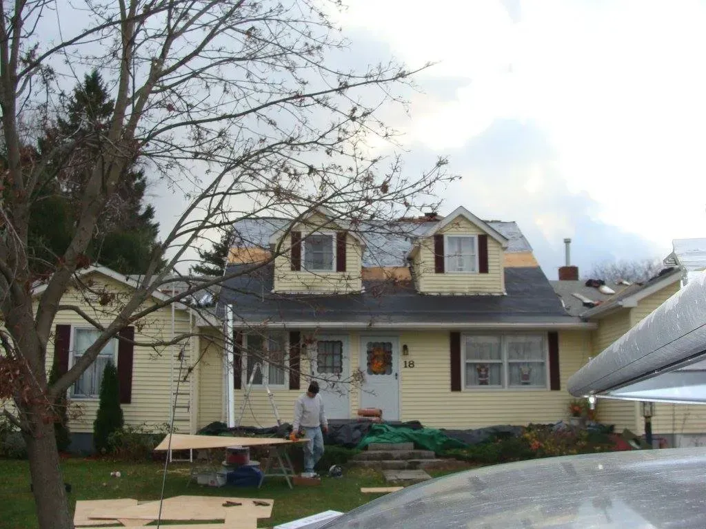 House with partial roof replacement; worker on the lawn near building materials; overcast sky.