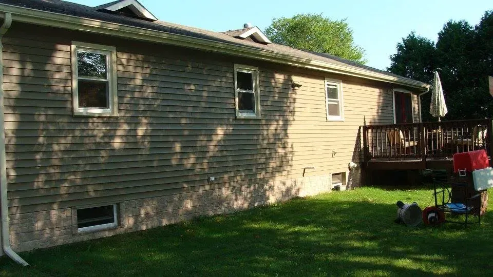 Side view of a house with tan siding and a small deck. Green grass in foreground.