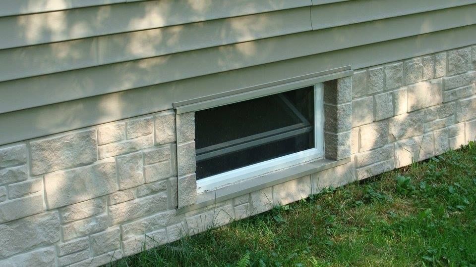 Window well on a house with stone-like siding, green grass, and gray siding above.