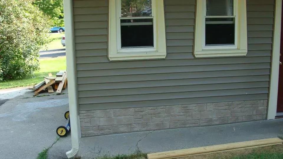 Gray house exterior with two windows and a brick-like lower section. A driveway, grass, and lumber are visible.