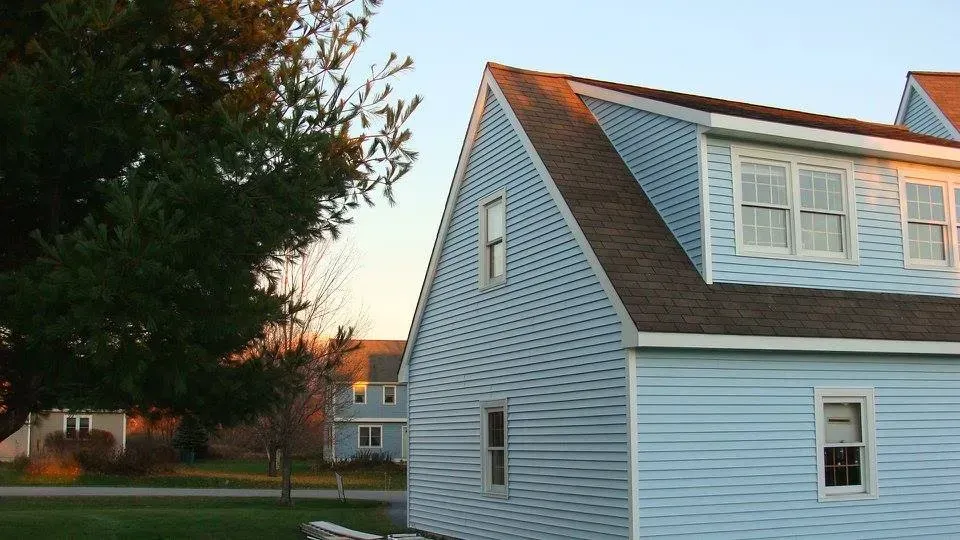 Blue house with dormers, brown roof, and small windows against a sunset sky; green lawn and tree in foreground.