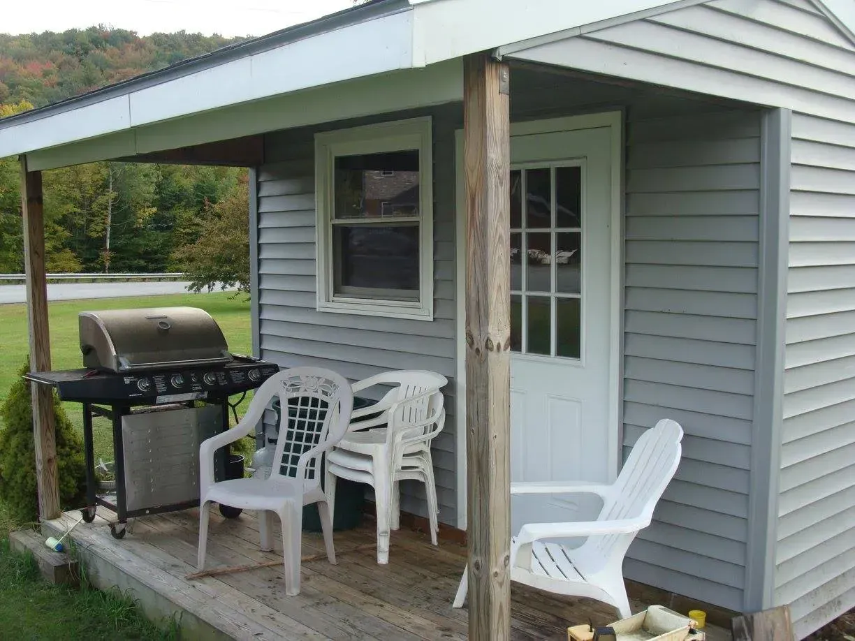 Small gray shed with a porch, grill, and white plastic chairs outdoors.