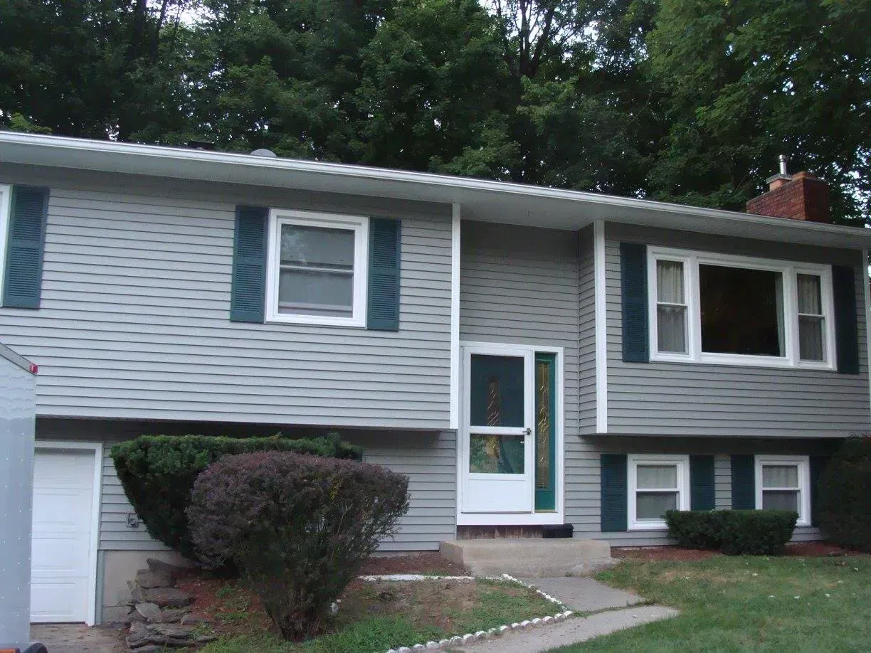 Gray two-story house with green shutters, white trim, and a brick chimney, set against a backdrop of trees.