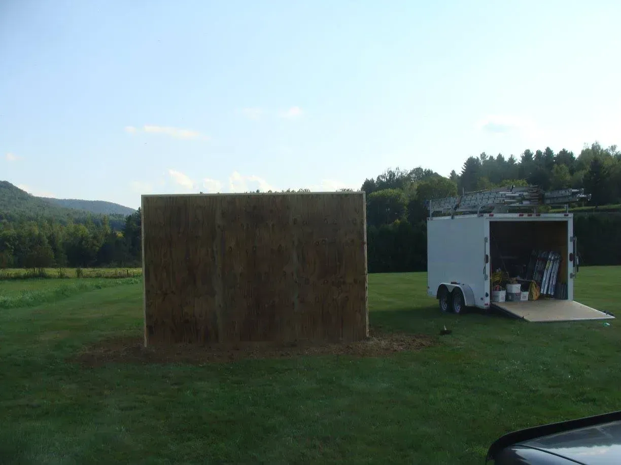 A large brown wall structure in a grassy field with a trailer next to it, mountains in the background.