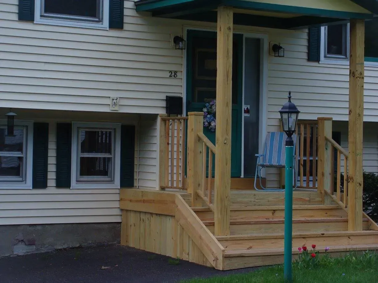 A wooden porch with steps and railing leads to a yellow house with a green roof and shutters.