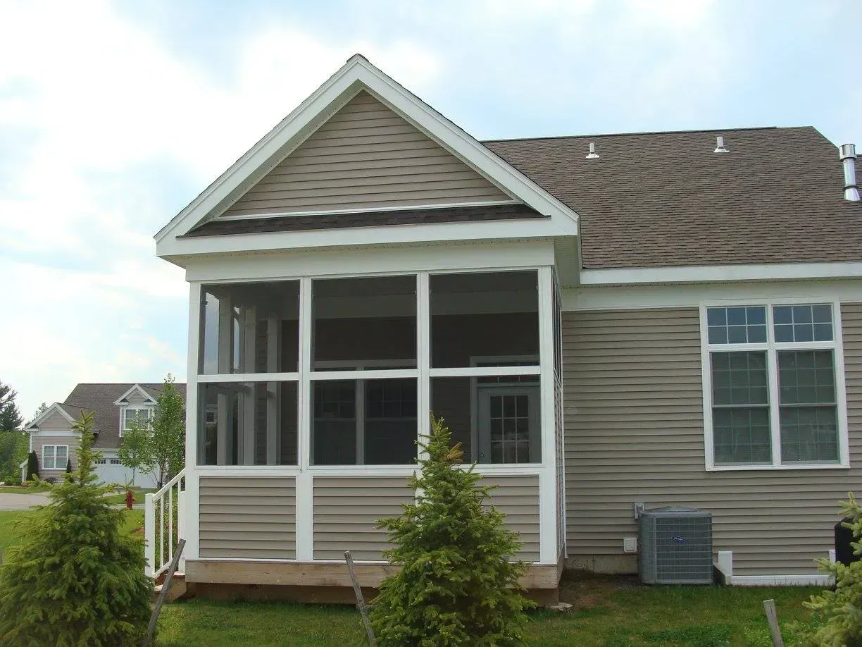 Screened-in porch attached to a house with tan siding and roof, two small evergreen trees in the foreground.